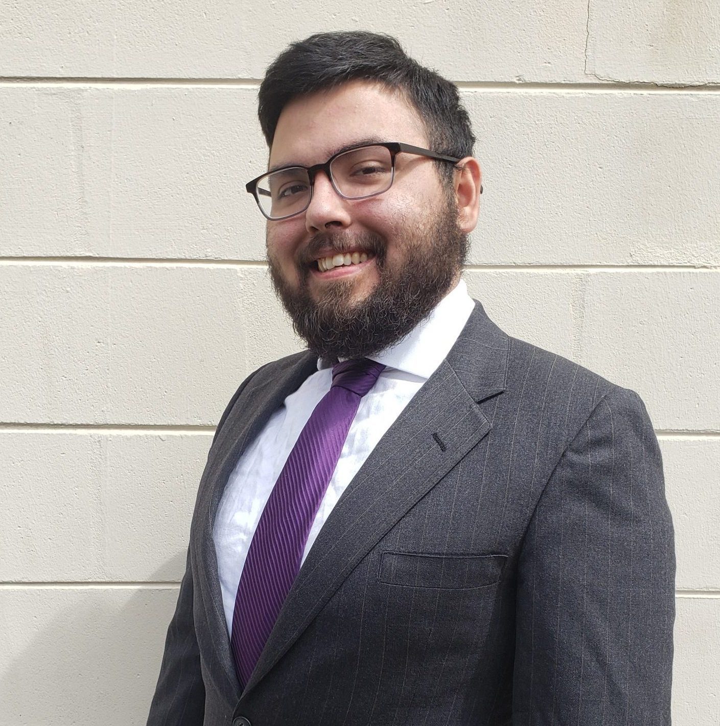 Jorge Alvarez in a black suit with a purple tie. Jorge stands in front of a white wall.